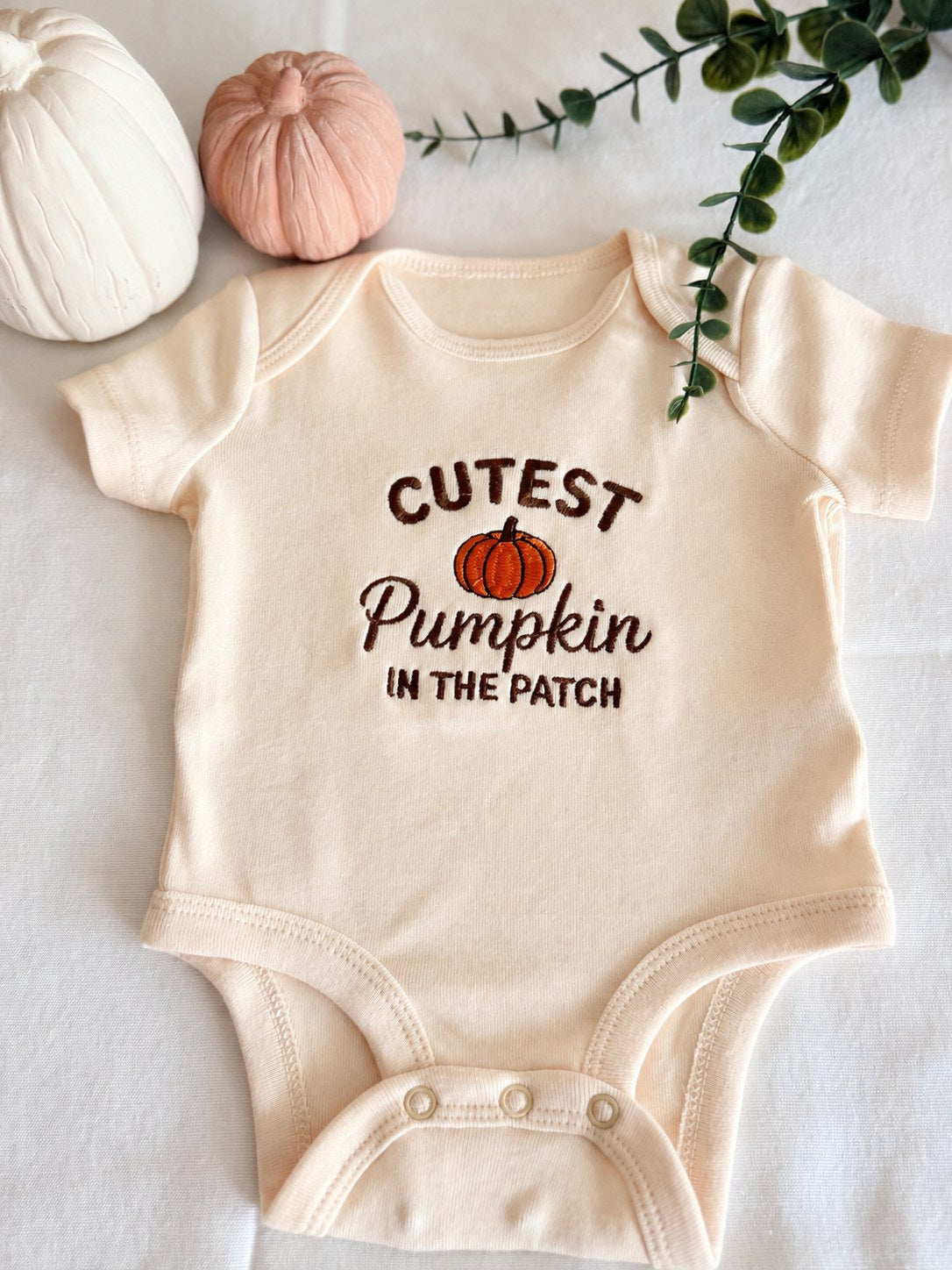 Sample: Cutest Pumpkin in the Patch embroidered on a natural baby bodysuit, with an orange pumpkin and brown lettering