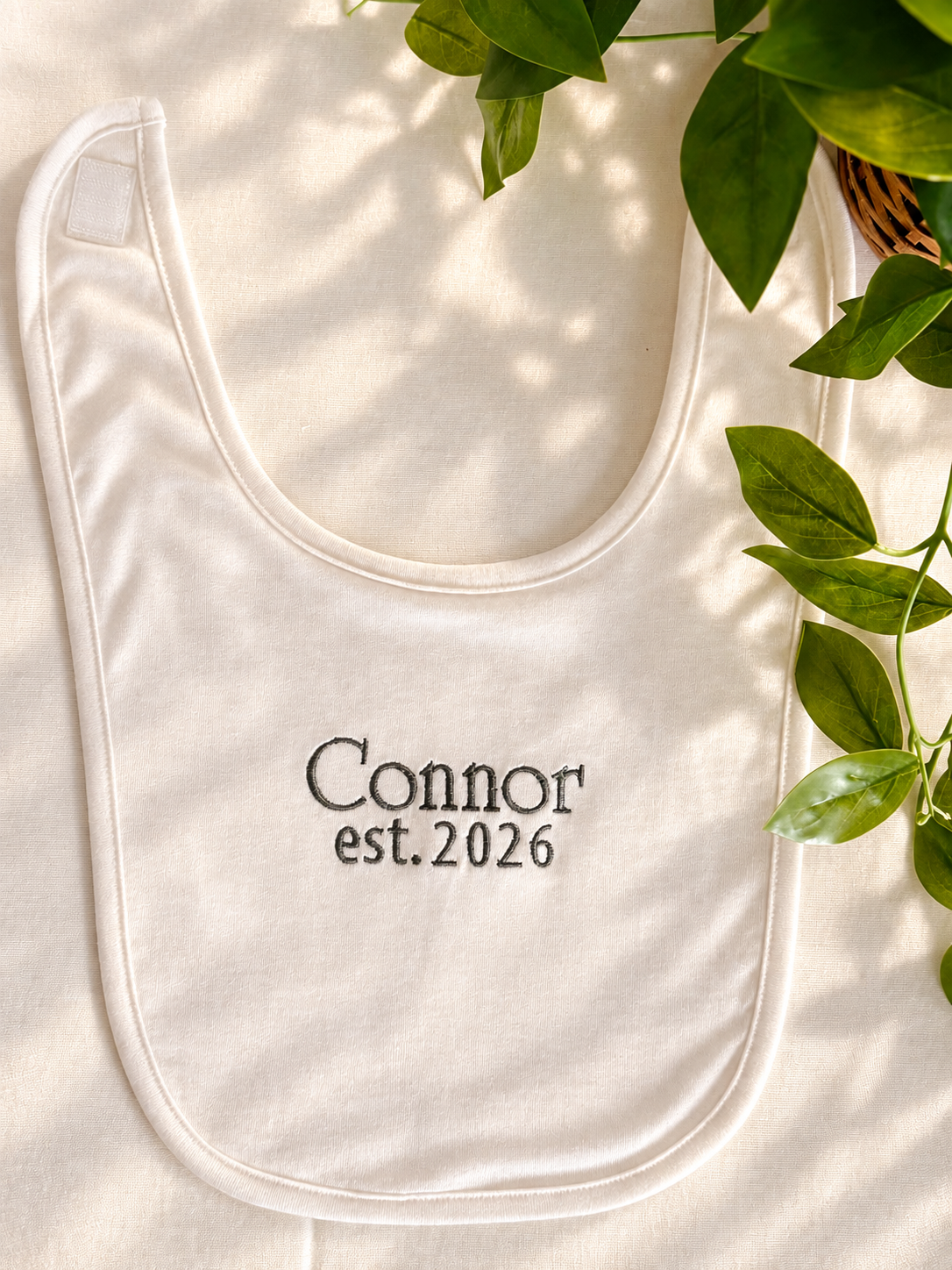 White baby bib embroidered with Connor and est. 2026 in dark gray thread, shown with green leaves.