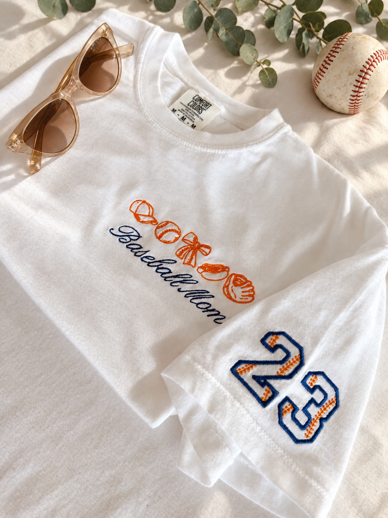 Sample: white Baseball Mom tee with navy “Baseball Mom” script and orange baseball icons on the left chest, and the number 23 embroidered on the sleeve