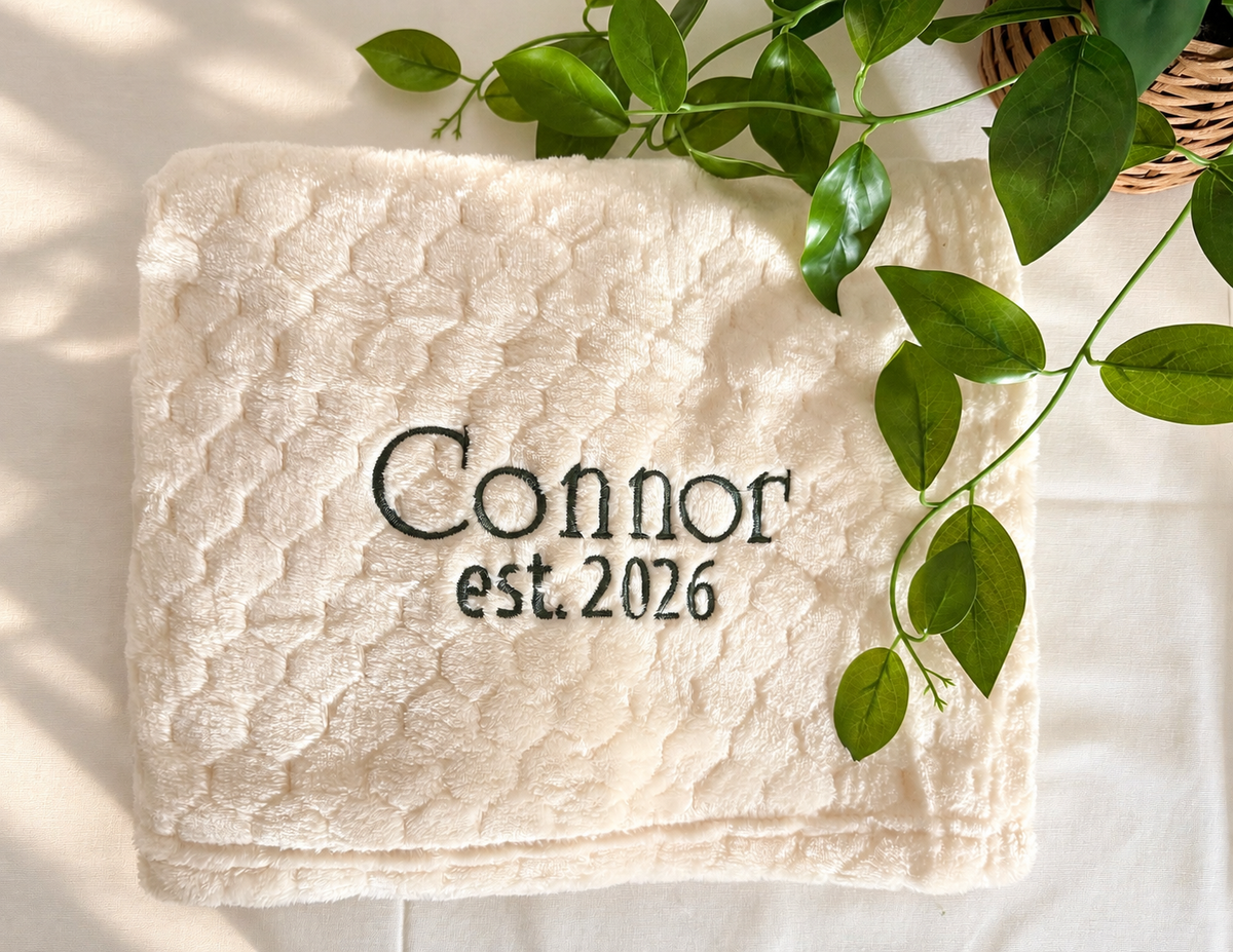 Soft cream honeycomb baby blanket embroidered with Connor and est. 2026 in dark green thread.