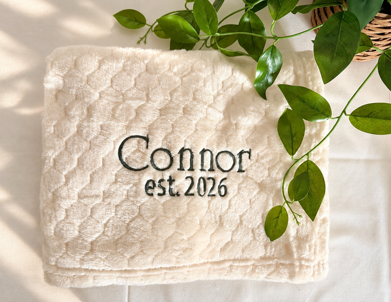 Soft cream honeycomb baby blanket embroidered with Connor and est. 2026 in dark green thread.