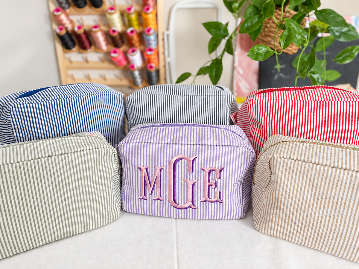Six striped seersucker cosmetic bags in navy, gray, red, sage, lavender and tan; the lavender bag shows an embroidered MGE monogram.