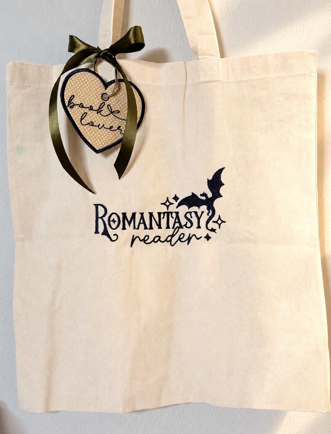 Sample: natural tote with Romantasy reader embroidered with a dragon and stars, and the heart book lover bag charm tied with an olive ribbon