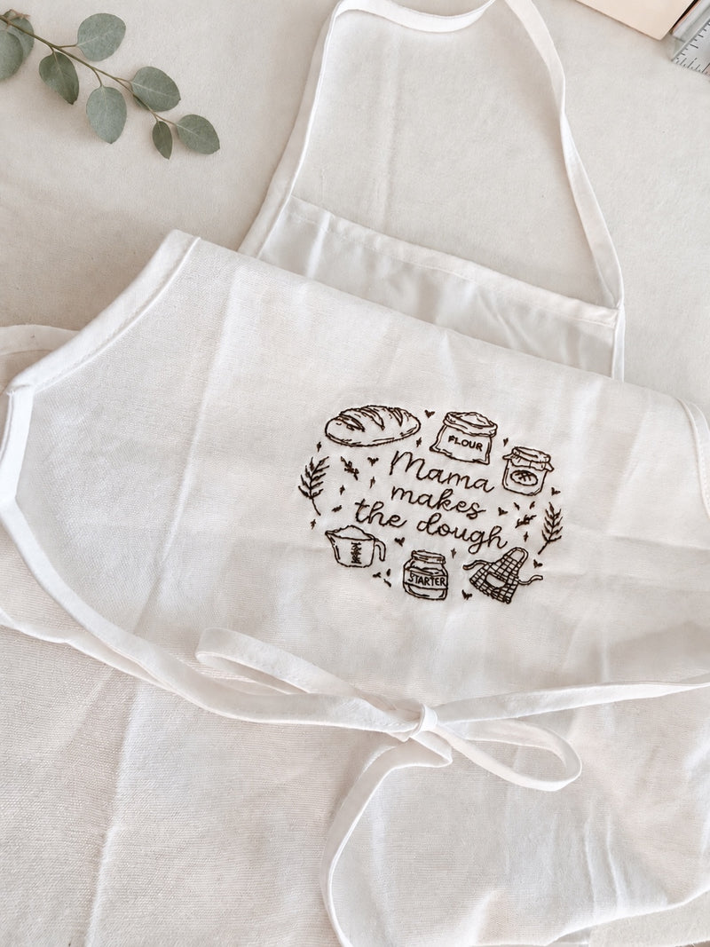 White apron with "Mama makes the dough" embroidered in brown script, surrounded by sourdough baking doodles