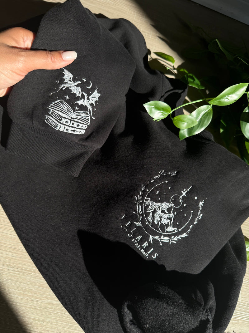 Sample Velaris sweatshirt laid flat, showing the embroidered chest design and the stacked books and dragons sleeve design